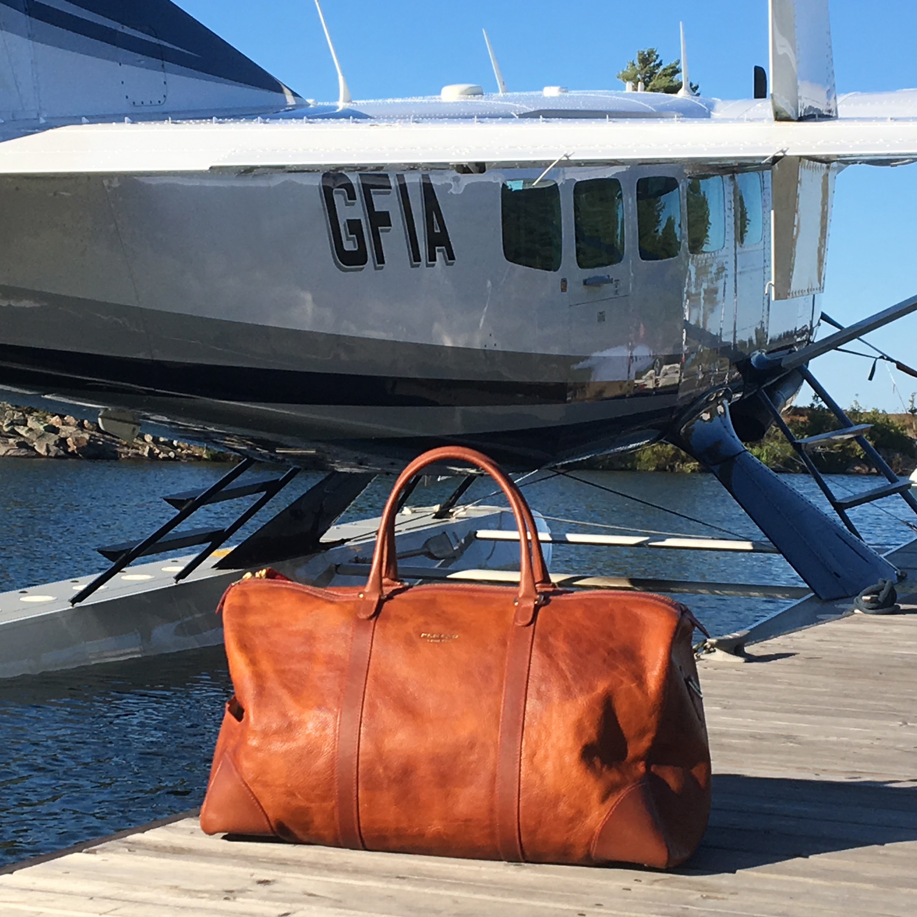Brown leather duffel bag on a dock with a seaplane in the background.