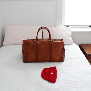 Brown leather bag and red hat on a white bed with a wooden nightstand.